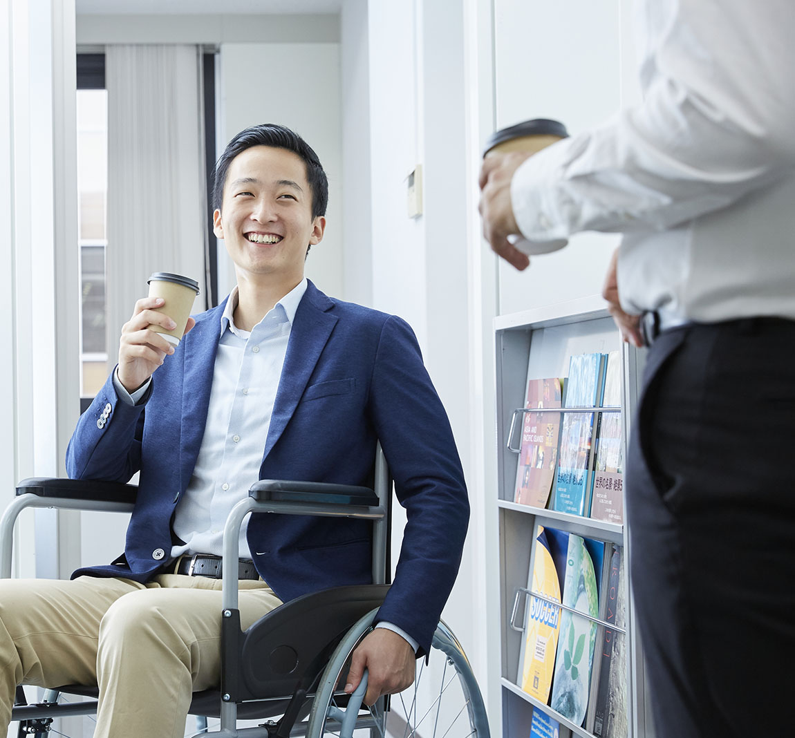 A man in a wheelchair holding a coffee cup smiles at a colleague.