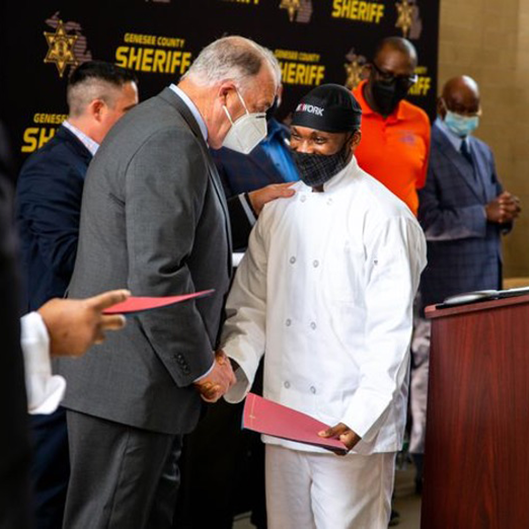 A suited official stands at a podium and shakes the hand of a smiling man wearing a chef's jacket and In2Work hat.